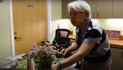 An older woman with short white hair, wearing a striped polo shirt, focuses intently on arranging a bouquet of flowers in a vase on a table. The arrangement features bright yellow daisies, pink blossoms, and small white flowers, creating a vibrant and delicate composition.